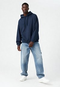 Námornícky modrá mikina s kapucňou a predným vreckom, skombinovaná so svetlomodrými džínsami a bielymi teniskami. Hladká tkanina a voľný strih.