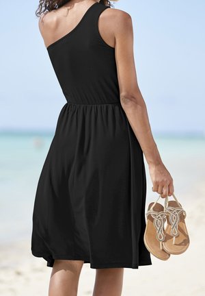 Czarna sukienka na jedno ramię z marszczonym pasem, zwiewny dół, trzymana przez osobę w opalonych sandałach z plecionym paskiem. Tło plażowe.