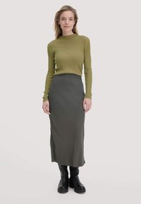 Grüner, gerippter, langärmeliger Oberteil mit hohem Kragen, kombiniert mit einem dunkelgrauen Midirock. Schwarze Boots mit dicker Sohle vervollständigen das Outfit.