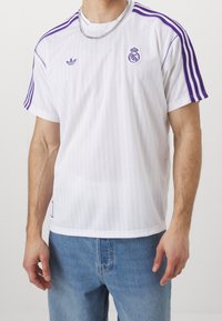 Vit träningsskjorta med vertikala ränder, lila detaljer, Adidas- och Real Madrid-logotyper på bröstet, korta ärmar och rundad halsringning.