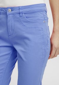 Svetle modré kalhoty s úzkým střihem, předními kapsami a kovovým zapínáním. Materiál má hladkou, měkkou strukturu s jemnými švy.