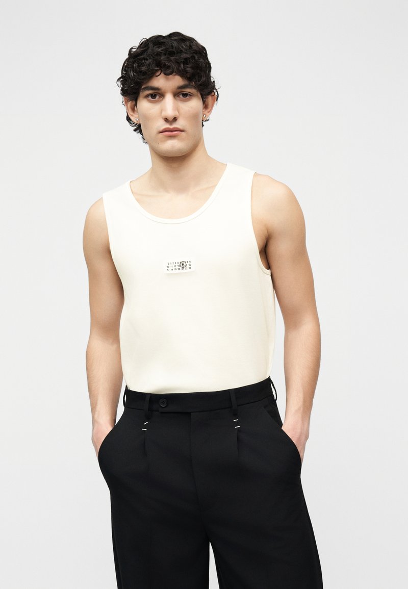 Giovane uomo con capelli ricci e scuri, indossa una canotta bianca e pantaloni neri, in piedi con le mani in tasca contro uno sfondo semplice.