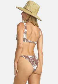 Hnedý bikini s bielym vzorom palmového listu, nastaviteľnými ramienkami a minimálnym pokrytím. Spárované s širokým slamenným klobúkom.