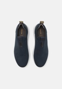 Jūras spēku tumši zili slip-on sporta apavi ar strukturētu adījuma augšdaļu, brūnu akcentu un balto zoli. Iezīmēti ar elpojošu materiālu un logo detaļām.