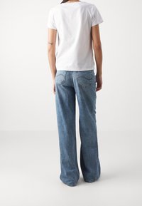 Personne portant un t-shirt blanc uni et un jean bleu à jambes larges, debout de dos face à la caméra sur un fond blanc.