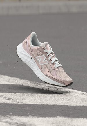 Chaussure de sport rose clair avec lacets et semelle blancs, flottant au-dessus d’un bitume avec des marquages blancs et petits débris noirs.