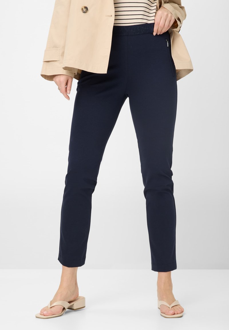 Marineblaue Slim-Fit-Hosen mit elastischem Bund und seitlicher Reißverschlusstasche, aus weichem Material; kombiniert mit hellen Sandalen und einer beigen Jacke.