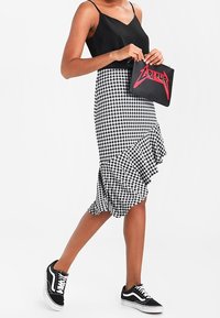 Kobieta nosząca czarny tank top, spódnicę w czarno-białą kratkę z falbankami, czarne sneakersy, trzymająca czarną kopertówkę z czerwoną grafiką.