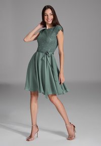 Kurzes grünes Kleid mit einem Spitzenoberteil, Pailletten und einem ausgestellten Rock. Verfügt über kurze Ärmel und einen gebundenen Taillengürtel. Glatte Textur.
