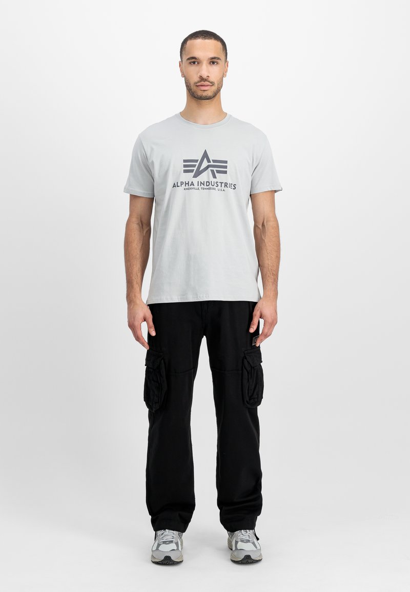 Férfi áll előre nézve, világosszürke Alpha Industries pólót, fekete cargo nadrágot és szürke sportcipőt viselve, fehér háttér előtt.