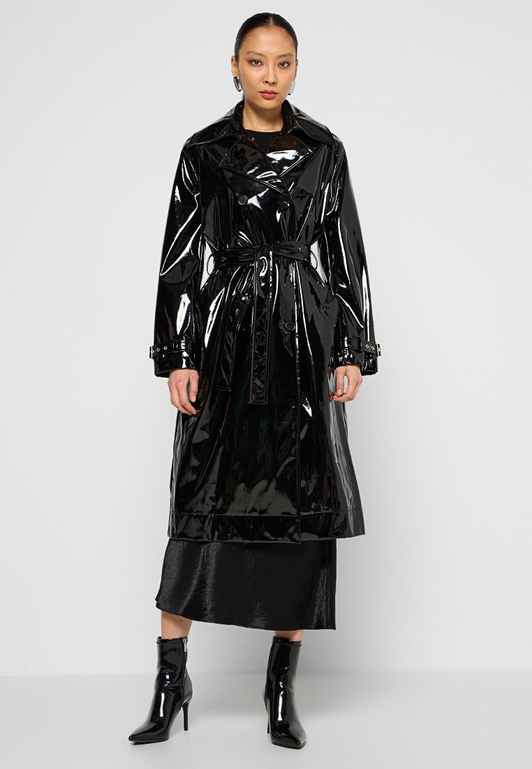 Schwarzer Lackleder-Trenchcoat mit tailliertem Schnitt, breiten Revers und geknöpften Manschetten, kombiniert mit einem schwarzen Satinkleid und Ankle Boots.