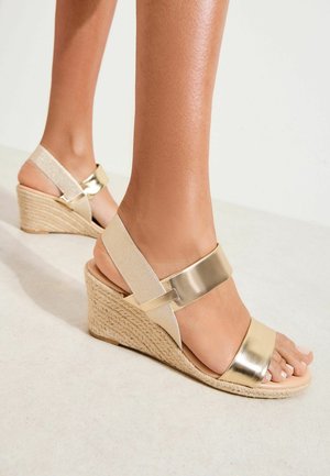 Jalat, joissa on beige-kultaiset kiiltonahkasandaalit punotulla köysikorolla, vaalealla pinnalla.