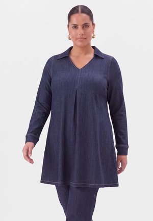 Femme aux cheveux foncés en chignon bas portant des boucles d'oreilles créoles dorées et une tunique bleu marine à manches longues avec un pantalon assorti sur un fond clair uni.