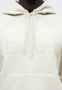 Φωτεινή μπεζ φουτέρ με κουκούλα από μαλακό βαμβάκι, με τσέπη καγκουρό, που διαθέτει ανάγλυφο λογότυπο "BOSS" στο μπροστινό μέρος και κορδόνια στη κουκούλα.