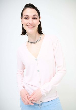 Giovane donna sorridente, indossa un cardigan rosa chiaro con bottoni, jeans azzurri e una collana argentata spessa, su uno sfondo bianco semplice.