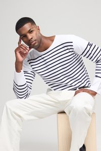 Langärmliges weißes T-Shirt mit navyblauen horizontalen Streifen, kombiniert mit crèmefarbenen Hosen, sitzt auf einem Holzstuhl vor einem hellen Hintergrund.