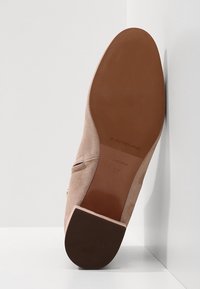 Zapato de ante beige con una puntera redondeada y suave, tacón bajo y un detalle en dorado. La suela es marrón con un acabado texturizado.