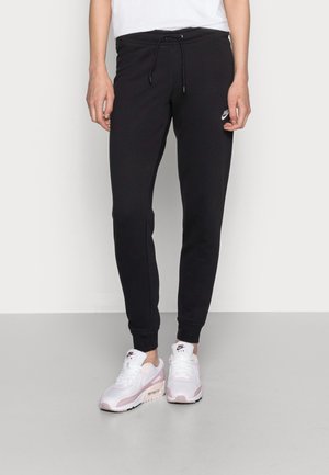 Person iført sorte Nike joggingbukser med snøre i taljen og hvide Nike Air sneakers med lyserøde detaljer, stående mod en ensfarvet baggrund.