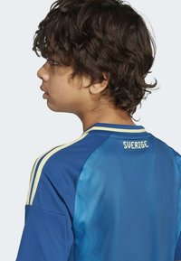 Modrý sportovní dres vyrobený z lehké tkaniny má žluté akcenty a na zadní straně text "SVERIGE". Krátké rukávy a design s kulatým výstřihem.