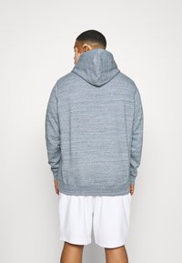 Felpa grigia con texture e cappuccio ampio, indossata sopra pantaloni corti bianchi, vista da dietro.