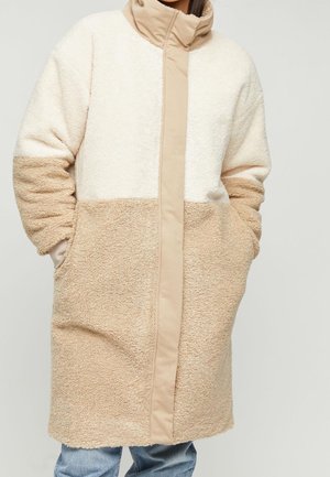 Femme portant un manteau sherpa polaire beige et crème à col montant, les mains dans les poches, associé à un jean bleu.