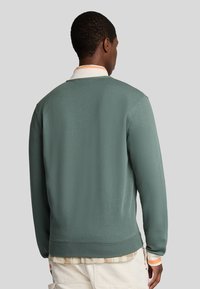 Sweatshirt vert avec une finition texturée, des poignets côtelés et un col rond, superposé sur une chemise à col avec des détails à carreaux au bas.