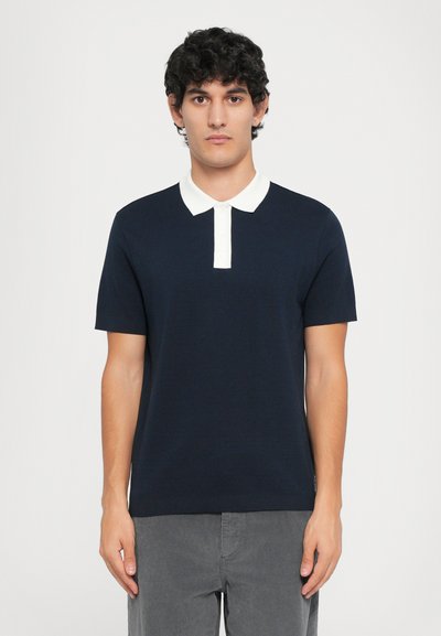 Polo shirt marine à manches courtes, col et patte de boutonnage blancs. Tissu texturé, coupe ajustée, associé à un pantalon en velours côtelé gris.