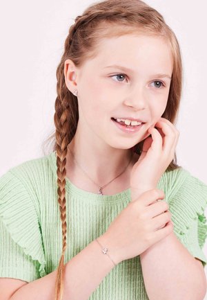 Silberschmuck mit einem kleinen Anhänger und mehrfarbigen Details. Das Modell trägt ein strukturiertes grünes Oberteil mit Rüschensäumen und hat eine geflochtene Frisur.