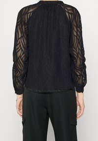 Blouse noire en tissu transparent avec un design texturé ondulé, manches longues, encolure froncée et poignets ajustés, associée à un pantalon noir ajusté.
