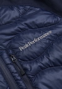 Navy blue insulated jacket with quilted texture, featuring a side zipper and embroidered "Peak Performance" logo in silver.