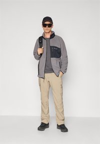 Giacca in pile grigia con tasca sul petto nera, t-shirt grigia, pantaloni cargo beige e scarpe da trekking nere, accessoriato con un cappellino e occhiali da sole.