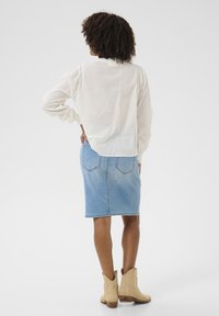 Camisa blanca de manga larga con hombros texturizados, combinada con una falda de mezclilla lápiz azul claro y botas vaqueras beige. Tejido suave, diseño casual.