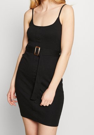 Vrouw die een aansluitende zwarte mouwloze jurk draagt met knopen aan de voorkant en een brede riem met een rechthoekige gesp in de taille.
