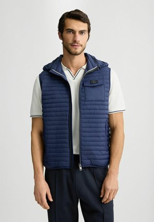 Dunkelblaues Puffer-Weste mit Kapuze, mit horizontalen Steppnähten und einer Reißverschlusstasche auf der Brust. Getragen über einem hellen Poloshirt.