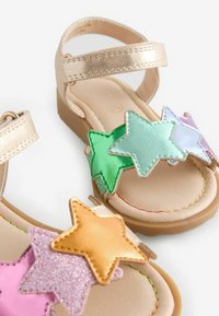 Gouden bandjes sandalen met glanzende sterdecoraties in roze, groen, paars en oranje. Zachte zool met een lichtbruine basis en gestikte details.
