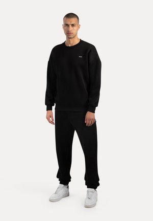 Felpa nera con collo rotondo, polsini a coste e un piccolo logo sul petto, abbinata a pantaloni della tuta neri coordinati e sneakers bianche.