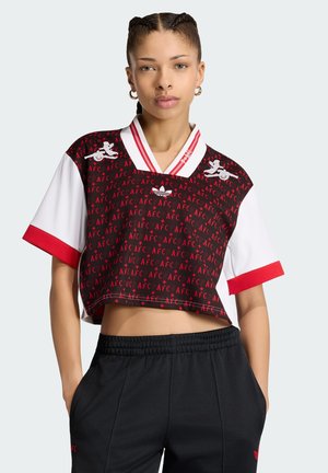 Junge Frau mit geflochtenem Haar, die ein kurzes schwarzes und rotes Oberteil mit AFC-Muster und schwarze Hose trägt, mit Händen in den Taschen stehend.