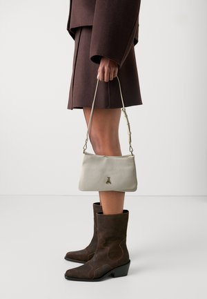 Wit leren handtas met een gouden accent, vastgehouden door een persoon in een donkerbruin oversized jasje en bruine suède enkellaarsjes met een blokhak.