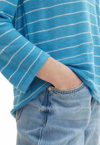 Blau-weiß gestreifter Pullover mit lockerer Passform, strukturiertem Strick und weiten Ärmeln, kombiniert mit hellblauen Jeans mit sichtbaren Knopfdetails.