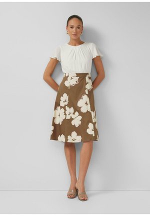 Femme portant un chemisier blanc à manches courtes et une jupe évasée marron avec de grandes fleurs blanches, debout contre un mur gris.