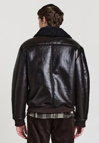 Braune Lederjacke mit strukturiertem Finish, ausgestattet mit einem schwarzen Schaffellkragen und gerippten Bündchen. Die Rückansicht zeigt deutliche Nahtlinien.