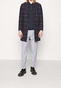 Manteau à carreaux bleu marine avec des accents rouges, porté sur un polo foncé, associé à un pantalon gris clair et des bottines noires à lacets. Textures lisses.