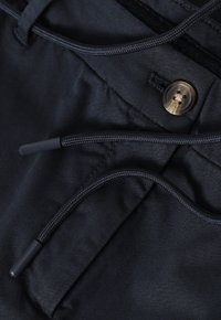 Pantalon marine avec une texture lisse, doté d'une fermeture à cordon et d'un bouton rond marron comme attache. Comprend des détails cousus.