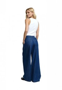 Pantaloni in denim a gamba larga di colore blu scuro con una finitura testurizzata, dotati di vita alta, passanti per cintura e due tasche posteriori. Abbinati a una canottiera bianca.
