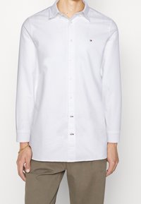 Camisa de manga larga blanca con cuello abotonado, tejido texturizado y pequeño logotipo en el lado izquierdo del pecho. Combinada con pantalones beige.