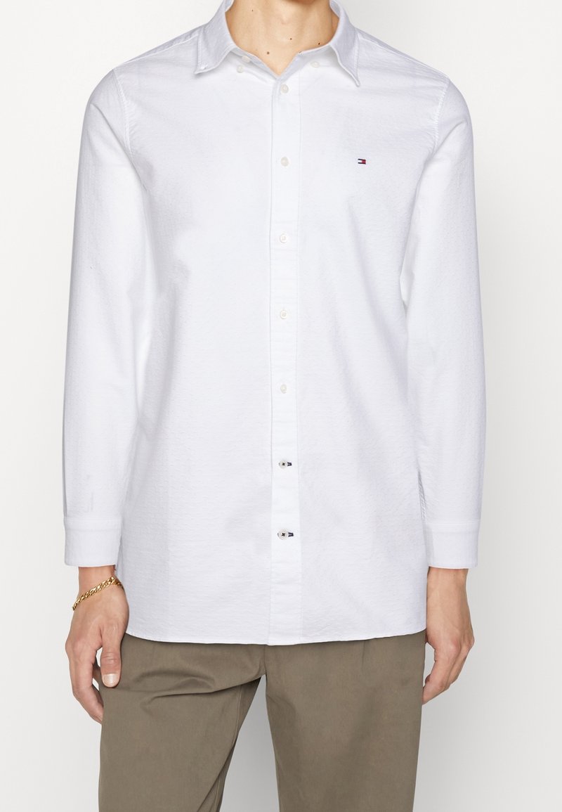 Camisa de manga larga blanca con cuello abotonado, tejido texturizado y pequeño logotipo en el lado izquierdo del pecho. Combinada con pantalones beige.
