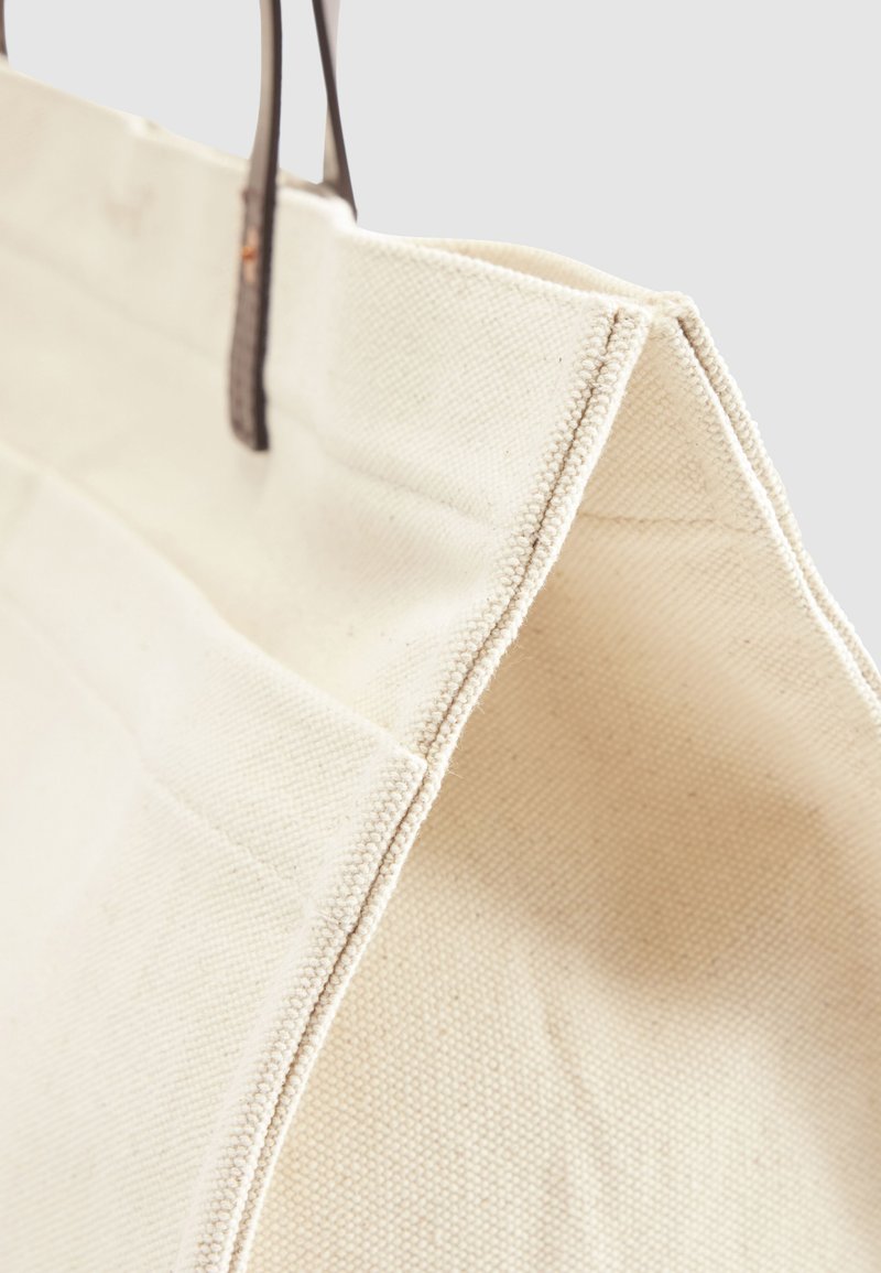Leinentasche in Cremeton mit dunkelbraunen Ledergriffen, Detailaufnahme der Seitennaht und Stoffstruktur.