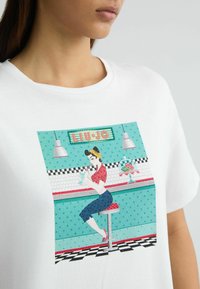 Balta T-shirt su spalvingu retro diner's atspaudų, vaizduojančiu moterį prie baro, su aqua fono ir taškeliais, raudonais akcentais ir juodais šaškių langeliais.