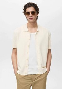 Cardigan corto crema a maniche corte con un tessuto testurizzato, chiusura con bottoni e colletto piatto, indossato sopra una canotta bianca e abbinato a pantaloni khaki.