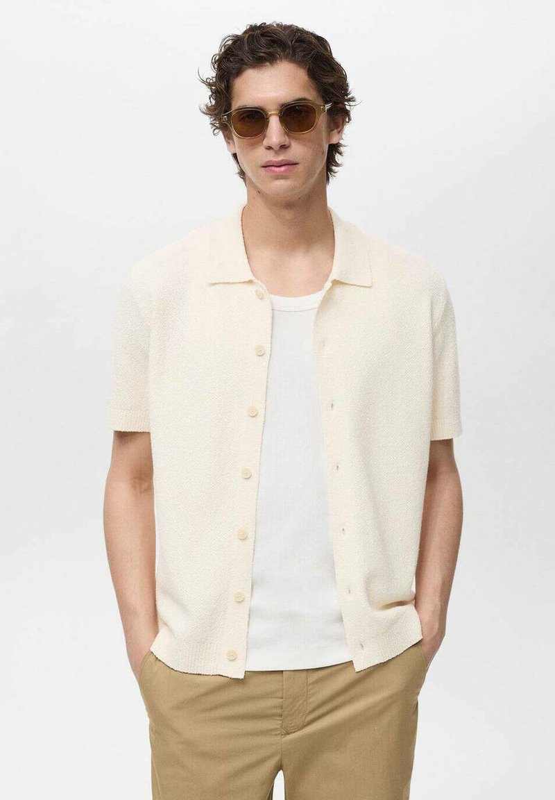 Cardigan corto crema a maniche corte con un tessuto testurizzato, chiusura con bottoni e colletto piatto, indossato sopra una canotta bianca e abbinato a pantaloni khaki.